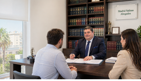 Abogado reunido con dos clientes en despacho jurídico con librería legal al fondo.