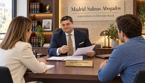 Juan Madrid Salinas, con traje azul marino, atiende a dos clientes en un despacho con rótulo de especialista en Derecho fiscal.